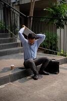 ett arg asiatisk affärsman sitter på utomhus- steg, innehav en bärbar dator med frustration, varelse avskedad. foto
