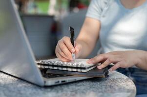 en närbild skott av en kvinna skrivning något i henne anteckningsbok på ett utomhus- tabell, arbetssätt avlägset. foto