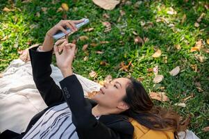 en Lycklig asiatisk affärskvinna är använder sig av henne smartphone medan liggande på en picknick matta i en parkera. foto