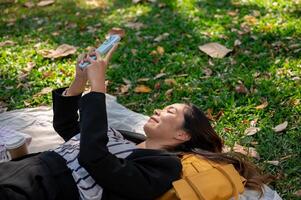 en Lycklig asiatisk affärskvinna är använder sig av henne smartphone medan liggande på en picknick matta i en parkera. foto