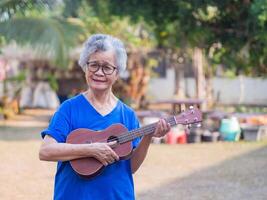 glad äldre asiatisk kvinna spelar de ukulele medan stående i de trädgård. Plats för text. begrepp av åldrig människor och avslappning foto