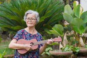 glad en senior kvinna spelar de ukulele medan stående i en trädgård. avkopplande förbi sång och spela små gitarr Lycklig och njut av liv efter går i pension. begrepp av åldrig människor och sjukvård foto