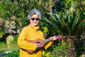 glad äldre asiatisk kvinna med kort grå hår bär glasögon och spelar de ukulele medan stående i en trädgård. begrepp av åldrig människor och avslappning foto