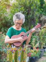porträtt av ett äldre asiatisk kvinna spelar de ukulele medan stående i en trädgård. Plats för text. begrepp av åldrig människor och avslappning foto