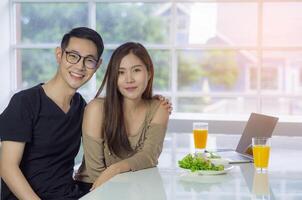 asiatisk ung par Sammanträde i de kök och ser på de kamera bredvid ha två glas av färsk orange juice, grönsaker sallad, bärbar dator och smartphone placerad på de tabell. par leende Lycklig foto