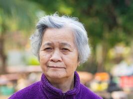 porträtt av äldre asiatisk kvinna med kort grå hår och stående leende och ser på de kamera medan stående i en trädgård. Plats för text. begrepp av åldrig människor och avslappning foto
