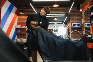 närbild av bearbeta av trimning av hår i barberare affär. kvalificerad barberare förvaring klippare i händer och korrigera form av hår till manlig klient Sammanträde på stol. begrepp av frisyr och rakning. foto