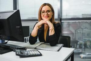 Söt, trevlig, söt, perfekt kvinna Sammanträde på henne skrivbord på läder stol i arbete station, bär glasögon, formell klädsel, har bärbar dator och anteckningsbok på de tabell foto