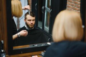 eleganta man Sammanträde barberare affär hårstylist frisör kvinna skärande hans hår porträtt stilig Lycklig ung skäggig caucasian kille få trendig frisyr attraktiv barberare flicka arbetssätt tjänande klient. foto