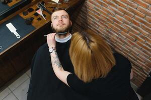skicklig kvinna barberare skärande en skägg av brunett vuxen kille i professionell retro frisör. foto
