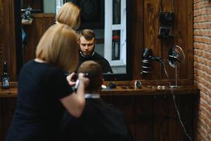 eleganta man Sammanträde barberare affär hårstylist frisör kvinna skärande hans hår porträtt stilig Lycklig ung skäggig caucasian kille få trendig frisyr attraktiv barberare flicka arbetssätt tjänande klient. foto