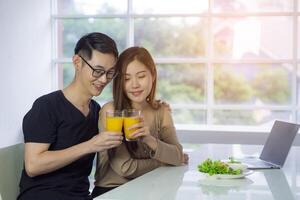 asiatisk ung par Sammanträde i de kök och innehav en glas av färsk orange juice för hälsa med vegetabiliska sallad en bärbar dator och smartphone placerad på de tabell. en par Lycklig är en Bra hälsa foto