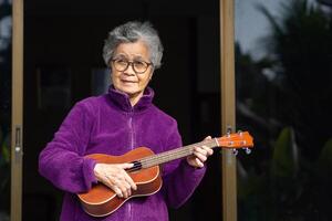 glad äldre asiatisk kvinna med kort grå hår bär glasögon och spelar de ukulele medan stående i främre av de dörr. begrepp av åldrig människor och avslappning foto