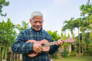 porträtt av en senior man leende och spelar de ukulele medan stående i en trädgård. Plats för text. begrepp av åldrig människor och avslappning foto