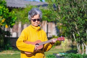 porträtt av en senior kvinna med kort grå hår spelar de ukulele, leende, och ser på de kamera medan stående i en trädgård. begrepp av åldrig människor och avslappning foto