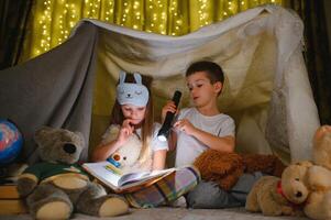 två barn med ficklampa läsa en bok under en filt som en tält foto