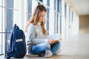 främre se av liten skön skola flicka bland korridor på skola, innehav anteckningar på händer. rolig och Lycklig flicka leende på kamera, vilar efter lektioner på primär skola foto