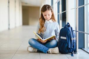 liten Söt flicka Sammanträde på de golv av de skola hall och läsning en bok. de begrepp av skolutbildning. foto