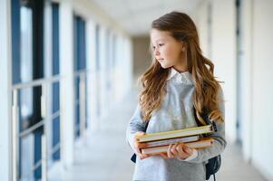 främre se av liten skön skola flicka bland korridor på skola, innehav anteckningar på händer. rolig och Lycklig flicka leende på kamera, vilar efter lektioner på primär skola foto