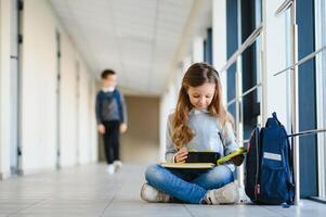 främre se av liten skön skola flicka bland korridor på skola, innehav anteckningar på händer. rolig och Lycklig flicka leende på kamera, vilar efter lektioner på primär skola foto