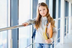 främre se av Söt blond skola flicka innehav många färgrik anteckningar och böcker. duktig tonåring flicka leende på kamera, stående på korridor av internationell skola. foto