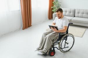 man i rullstol använder sig av läsplatta dator på Hem. positiv pensionerad manlig med fysisk handikapp bläddring webb på Rör vaddera, tittar på uppkopplad inomhus. foto
