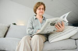 senior lady läsning henne tidning på Hem avkopplande på en soffa och peering över de topp på de visare foto