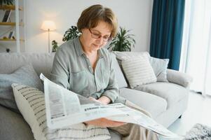 senior lady läsning henne tidning på Hem avkopplande på en soffa och peering över de topp på de visare foto