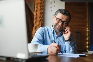 positiv senior skäggig man med grå hår dricka kaffe och använder sig av bärbar dator på Kafé, kopia Plats. eleganta åldrig affärsman i vinröd jacka njuter hans te medan arbetssätt uppkopplad. foto