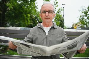 stilig farfar sitter på en bänk i de parkera och läser en tidning. senior gråhårig man. foto