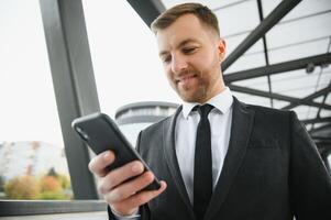Lycklig leende affärsman bär svart kostym och använder sig av modern smartphone nära kontor på tidigt morgon, framgångsrik arbetsgivare till göra en handla medan stående nära skyskrapa kontor på natt, blossa ljus foto