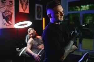 tatuerare i svart handskar drömmande ser i kamera innehav tatuering maskin i hand i modern tatuering studio foto