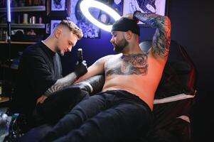 manlig tatuering konstnär innehav en tatuering pistol, som visar en bearbeta av framställning tatueringar på en manlig tatuerade modellens ärm foto