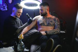 skäggig tatuering konstnär arbetssätt på hans studio tatuering ärm på de ärm av hans manlig klient. man få tatuerade förbi professionell tatuerare foto