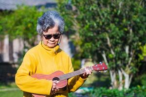 glad äldre asiatisk kvinna med kort grå hår bär solglasögon och spelar de ukulele medan stående i främre av de dörr. begrepp av åldrig människor och avslappning foto