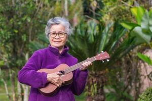 glad äldre asiatisk kvinna med kort grå hår bär glasögon och spelar de ukulele medan stående i en trädgård. begrepp av åldrig människor och avslappning foto