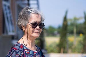 en porträtt av ett äldre kvinna bär solglasögon medan stående i en trädgård. Plats för text. begrepp av gammal människor och Semester. foto