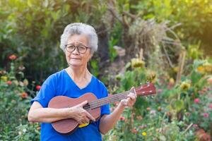 glad senior kvinna med kort grå hår spelar de ukulele, leende och ser på de kamera medan stående i en trädgård. begrepp av åldrig människor och avslappning foto
