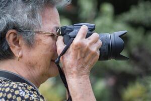 senior kvinna med kort grå hår bär glasögon och skytte fotografi förbi en digital kamera på de parkera. begrepp av åldrig människor och fotografi foto