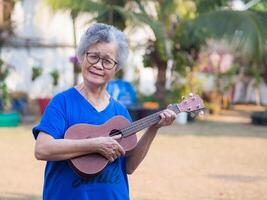 glad senior kvinna med kort grå hår spelar de ukulele, leende och ser på de kamera medan stående i en trädgård. begrepp av åldrig människor och avslappning foto