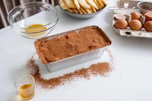 tiramisu framställning bearbeta. italiensk efterrätt i folie bricka och Ingredienser på kök tabell foto