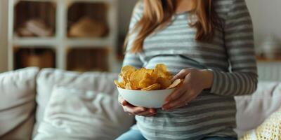 gravid kvinna Sammanträde på de soffa åtnjuter äter potatis pommes frites från en skål på Hem. foto