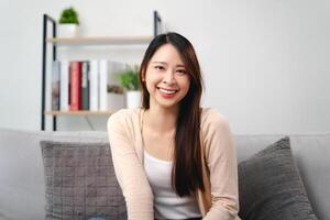 en porträtt av skön ung asiatisk kvinna med lång brun hår är Sammanträde på en soffa och leende foto