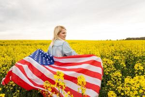 attraktiv kvinna innehav ett amerikan flagga i de vind i en fält av raps. sommar landskap mot de blå himmel. horisontell orientering. foto