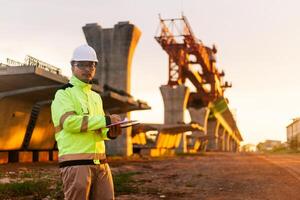en konstruktion arbetstagare är stående på en bro, talande på hans cell telefon. de scen är uppsättning i en konstruktion webbplats, med en stor kran i de bakgrund. de arbetstagare är bär en gul jacka foto