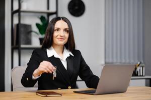 ung Lycklig anställd företag kvinna bär kostym Sammanträde på kontor skrivbord med bärbar dator och ger nyckel i kontor foto