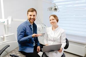 en stilig ung man är talande till en kvinna läkare på en dental utnämning i en ljus, skön kontor. de tandläkare förklarar till de patient och visar allt på de bärbar dator foto