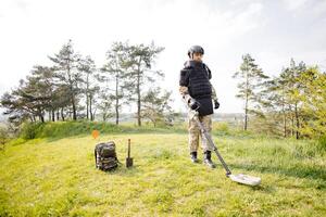 en man i en militär enhetlig och skottsäker väst Arbetar i de skog med en metall detektor. en minsvepare utför arbete på minröjning de territorium foto