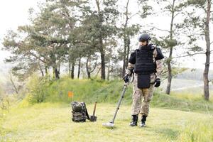 en man i en militär enhetlig och skottsäker väst Arbetar i de skog med en metall detektor. en minsvepare utför arbete på minröjning de territorium foto