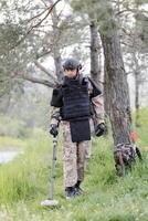 en man i en militär enhetlig och skottsäker väst Arbetar i de skog med en metall detektor. en minsvepare utför arbete på minröjning de territorium foto